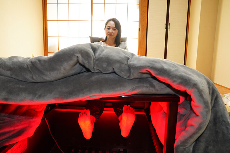 和室のこたつ下で赤く照らされた足先を見せ、奥に座る安野由美を捉えた場面