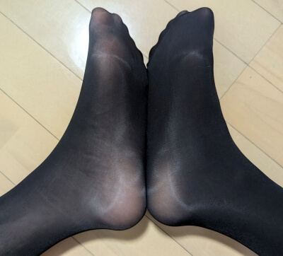 光沢パンストを履いた両足を揃え、照明を反射して艶めかしく輝く足元をアップで捉えている